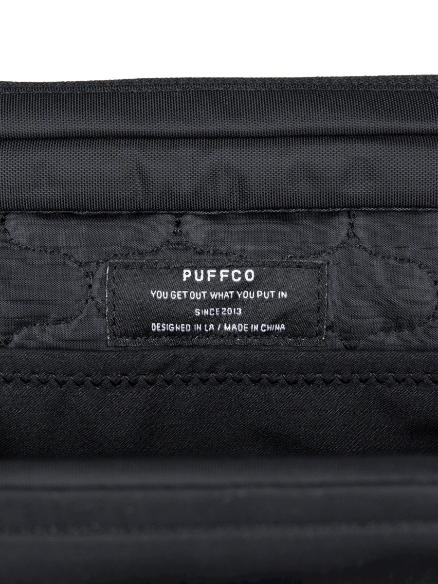 a close-up view of a black fabric, possibly a piece of clothing or a bag, with a label or tag attached to it. The label contains text that reads "PUFFCO" and "YOU GET OUT WHAT YOU PUT IN SINCE 2013 DESIGNED IN LA / MADE IN CHINA".