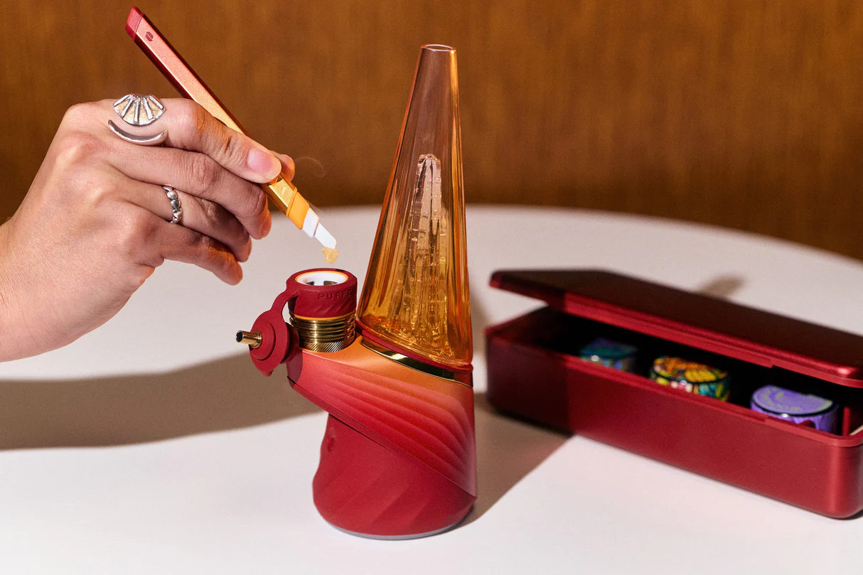 A hand is holding a red and gold pipe, with a red and gold pipe cleaner in the foreground, and a red and gold pipe in the background.