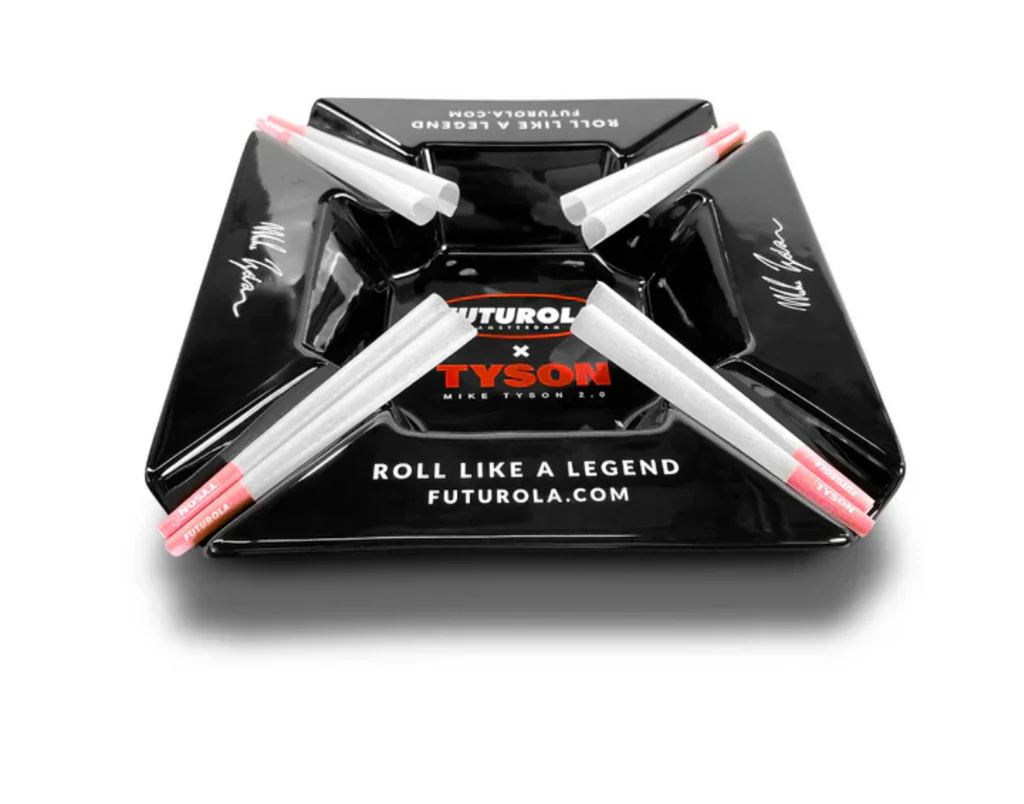 A black ashtray with the text "FUTUROLA.COM" and "TYSON" printed on it, along with the phrase "ROLL LIKE A LEGEND" and "MIKE TYSON 2.9" printed on it. There are also four white and pink cigarette butts arranged in a cross pattern on the ashtray.