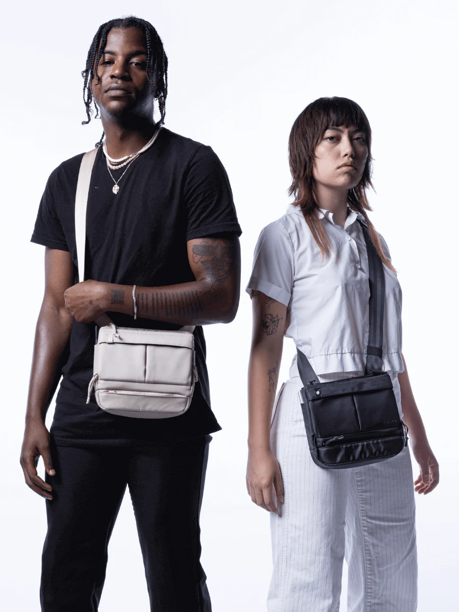A man and a woman standing side by side, both carrying bags.