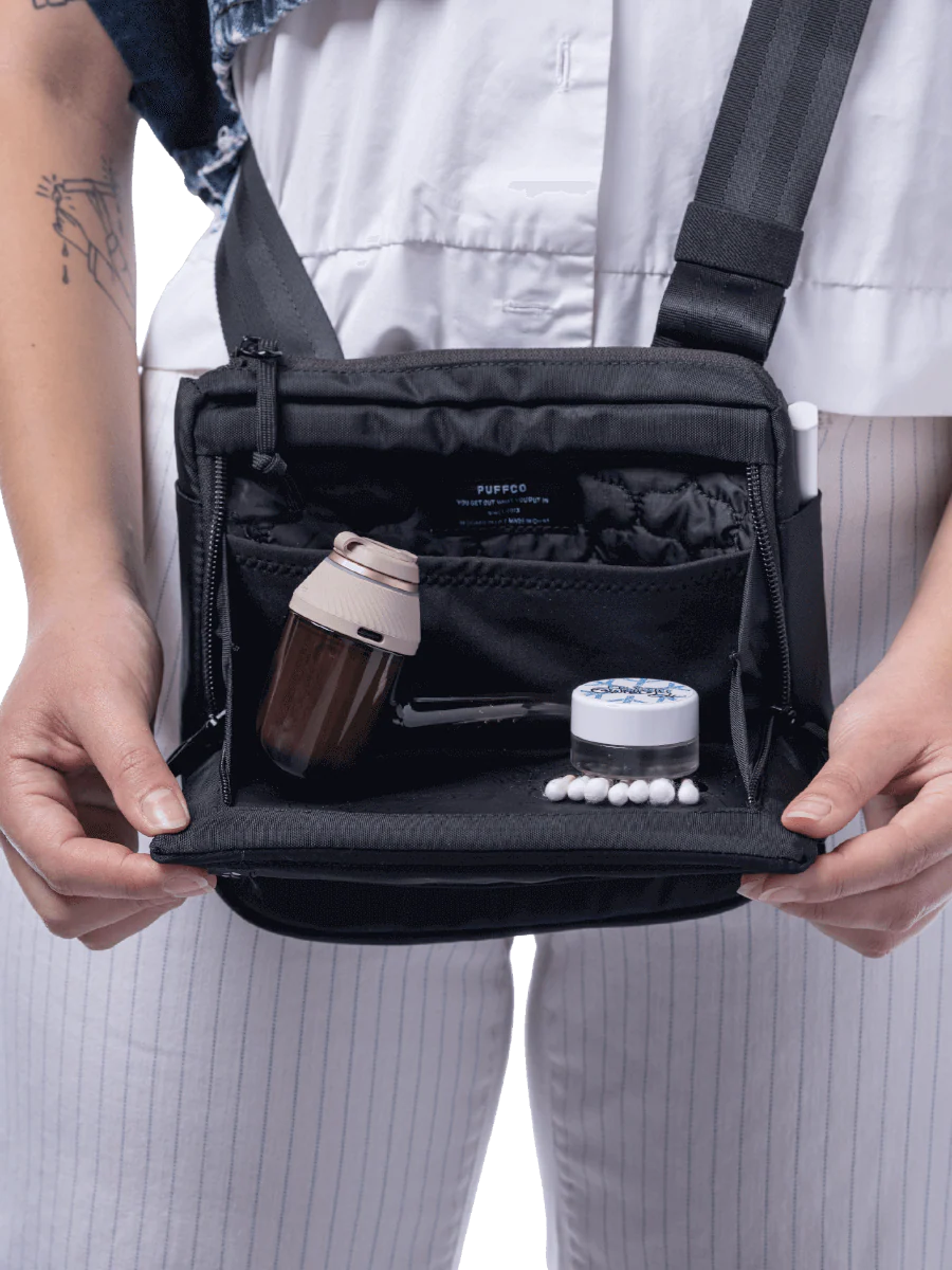 A person is holding a black bag with a small bottle and a tube of toothpaste visible inside.