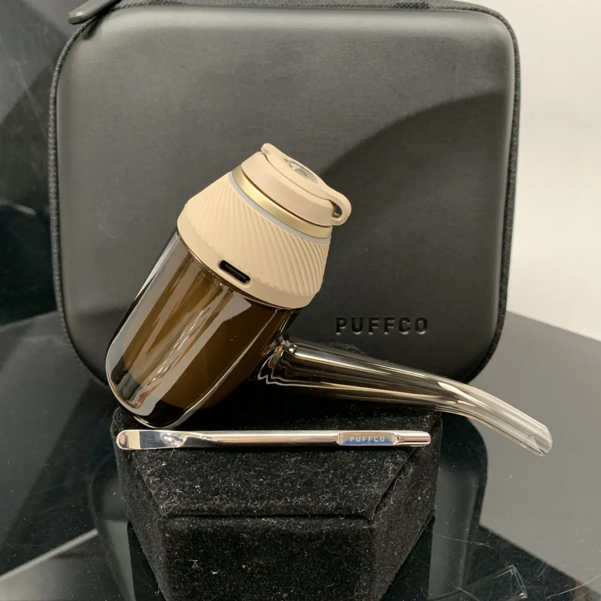 A brown glass pipe with a white lid and a black case with the word "PUFFCO" printed on it are displayed on a black surface.
